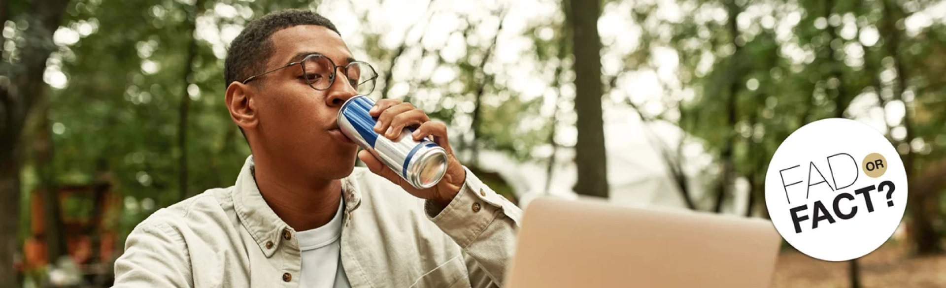 A young man has a sip of an energy drink while sitting outside in front of a laptop computer.