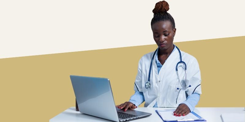 Person in white coat with stethoscope around neck, looking down at paper chart and entering information into laptop