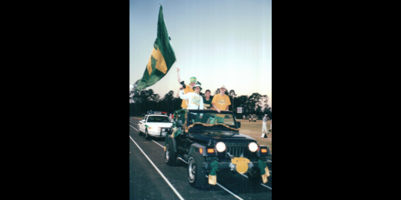 High schoolers riding in a jeep during a parade holding a flag