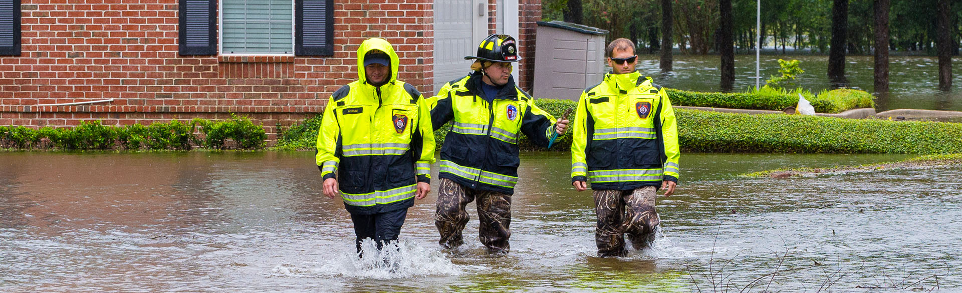 Three firefighters clad in black and bright yellow gear wade through knee-deep water while staying close together