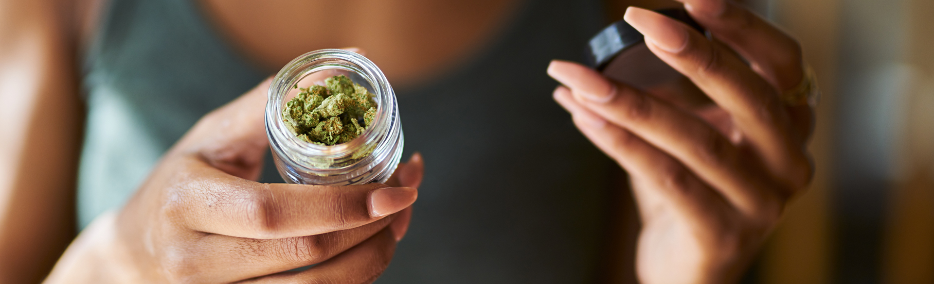 Woman holds a small jar of cannabis flower in one hand with lid in other hand
