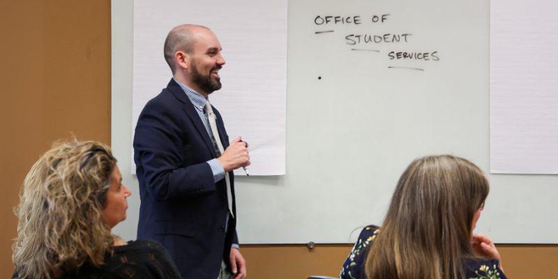 A man with a short beard and suit runs a meeting. 