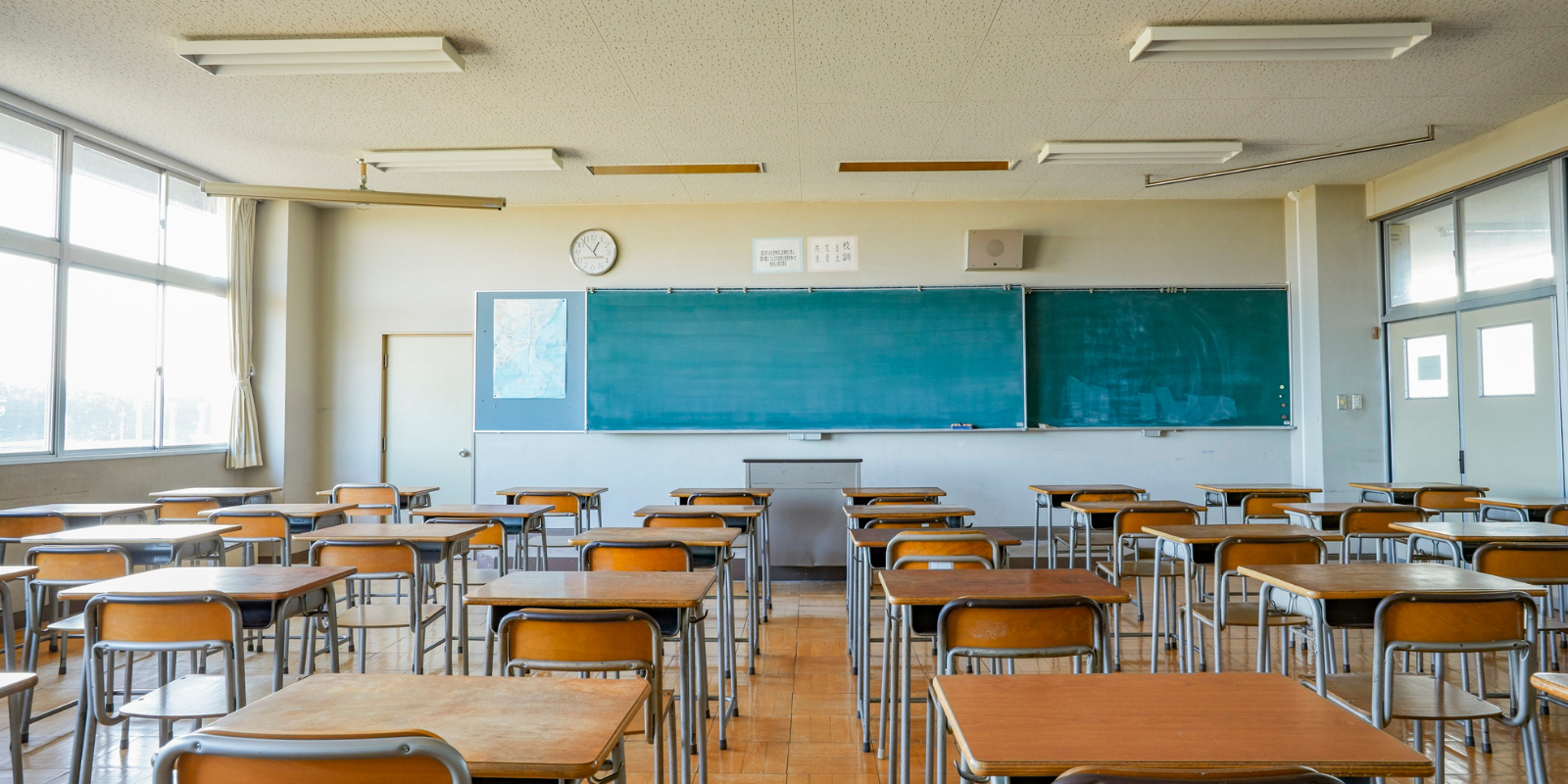 A bright, empty classroom with rows of wooden desks and orange chairs facing a large teal chalkboard, with windows along the left wall and fluorescent lighting overhead.