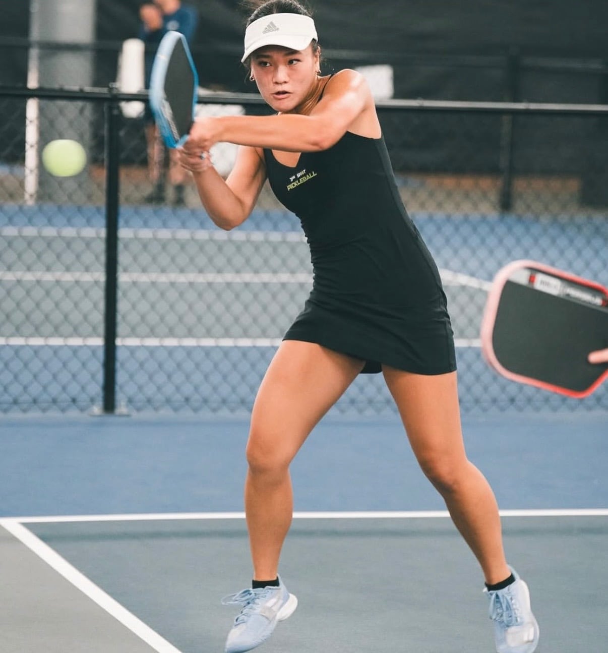 Eileen Tran playing pickleball, wearing black with a white hat, having just hit a ball
