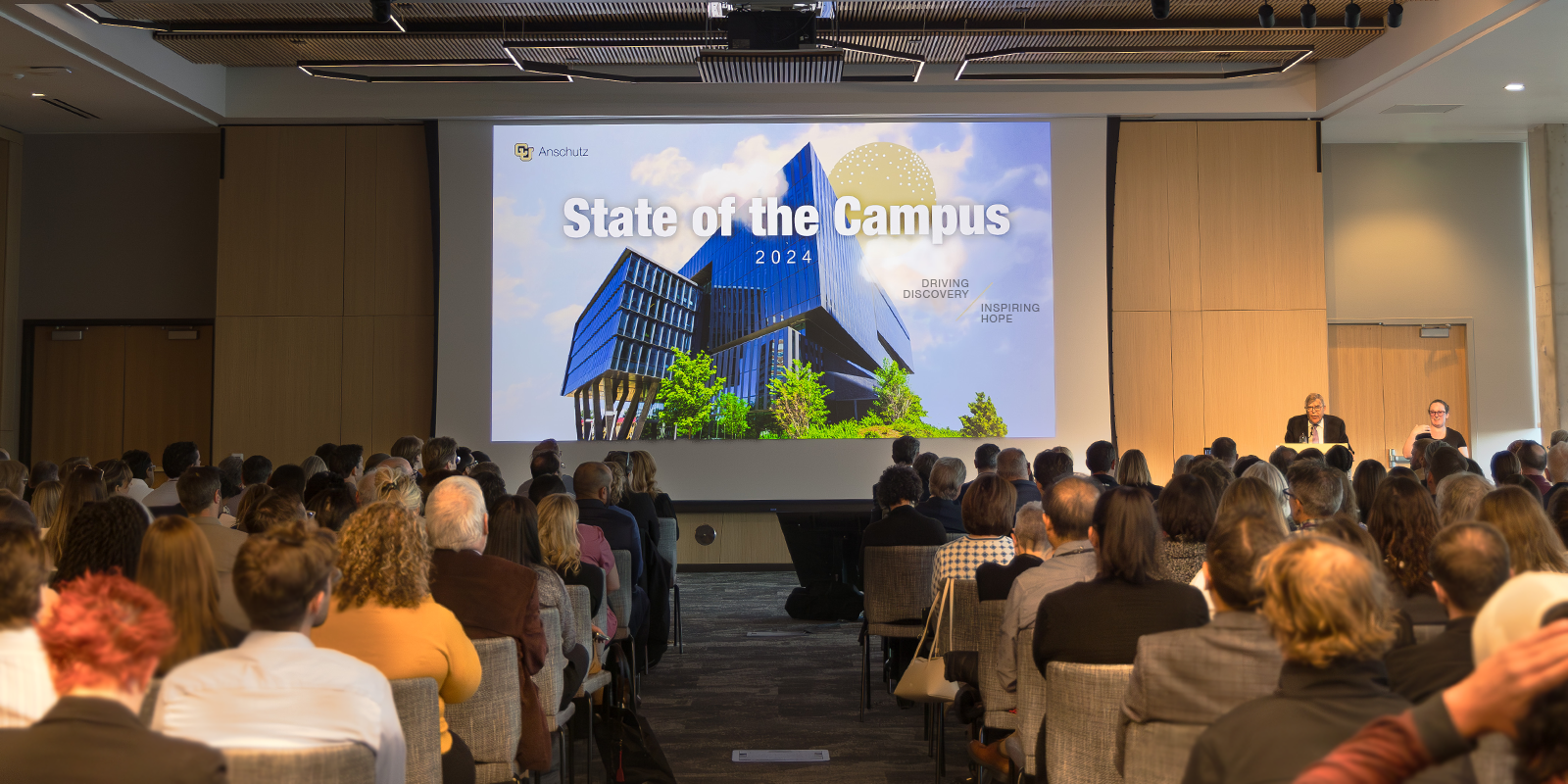 A photo from the back of the audience looking straight at the large screen with the Anschutz Health Sciences Building