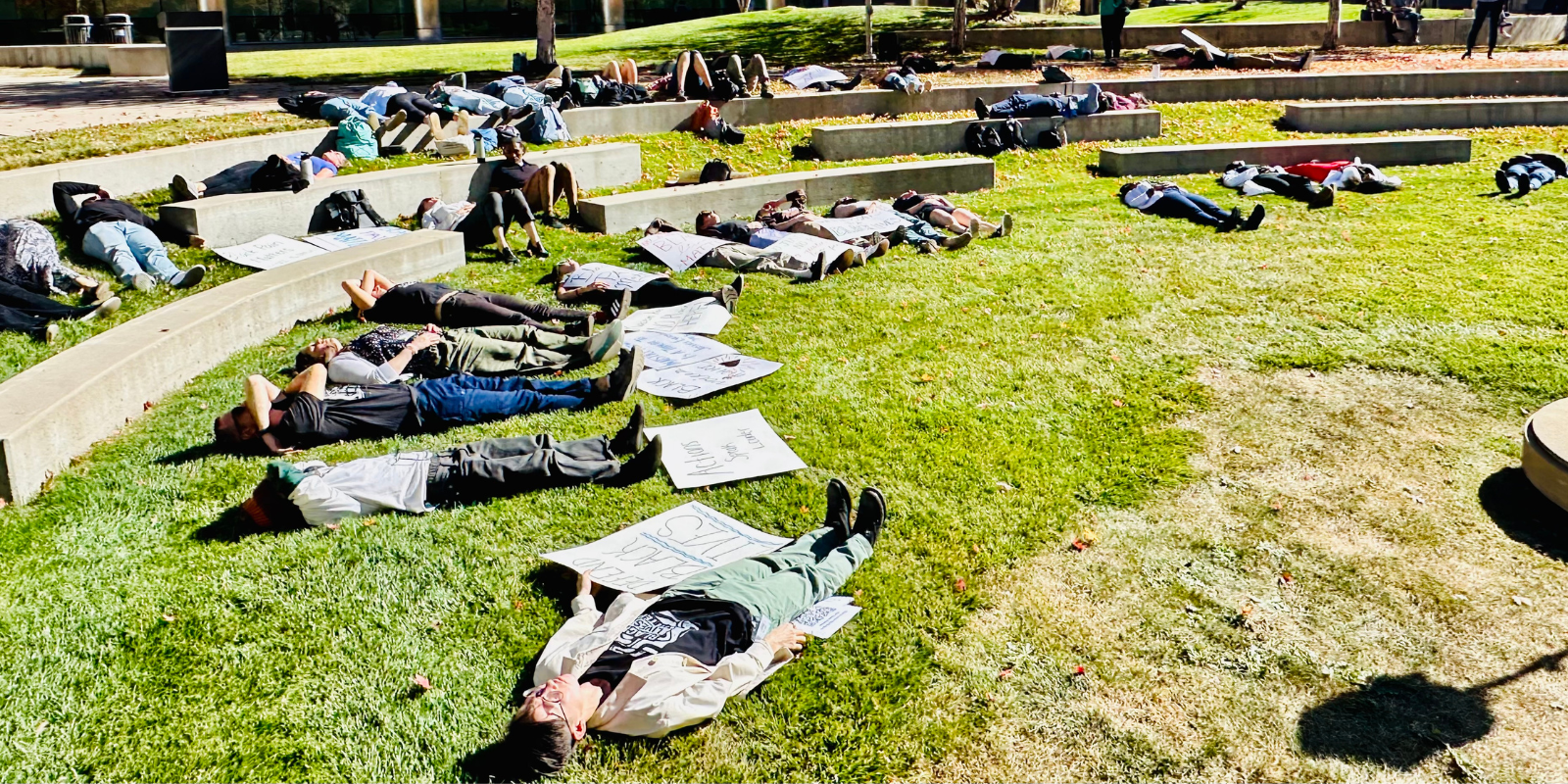 Community members lying on grass with signs beside them.