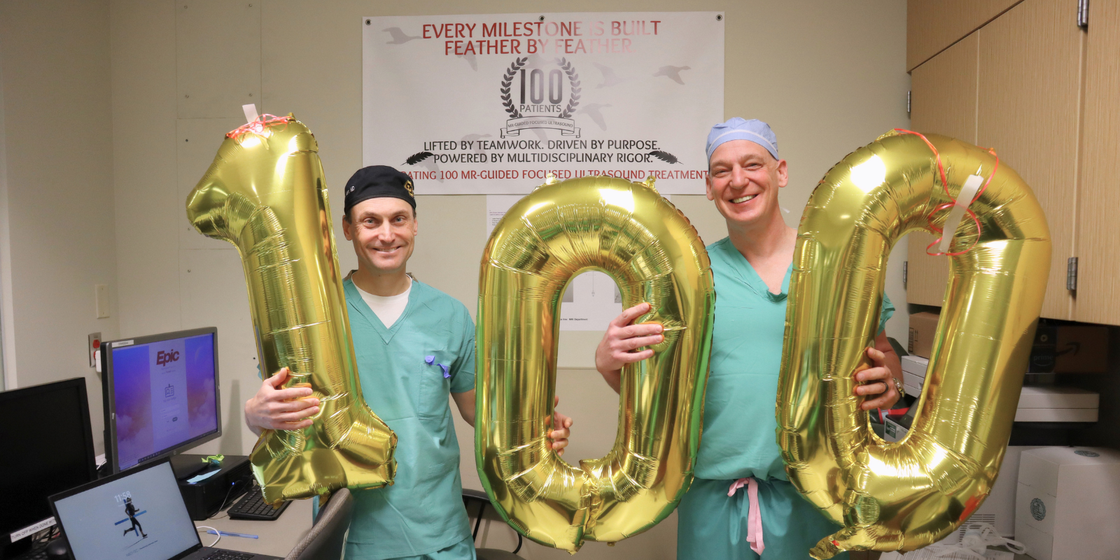 An image of two doctors dressed in blue scrubs holding up balloons that spell out 100.