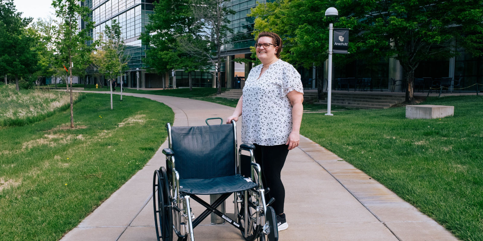 Lynn Montgomery-Haga stands next to an empy wheelchair on a grass-lined sidewalk on the CU Anschutz Medical campus