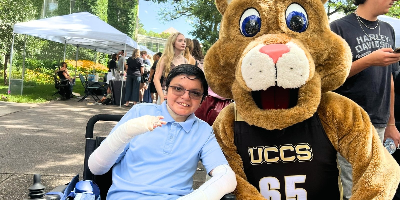 Harshit Talwar, his arms wrapped in white bandages, smiles as he poses in a wheelchair next to CU Colorado Springs mascot Clyde the mountain lion.