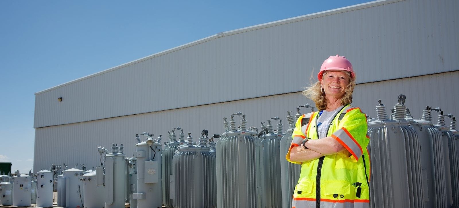 Construction worker, woman with blonde hair and safety equipment, stands in front of electrical machinery. Woman is smiling and has arms crossed.