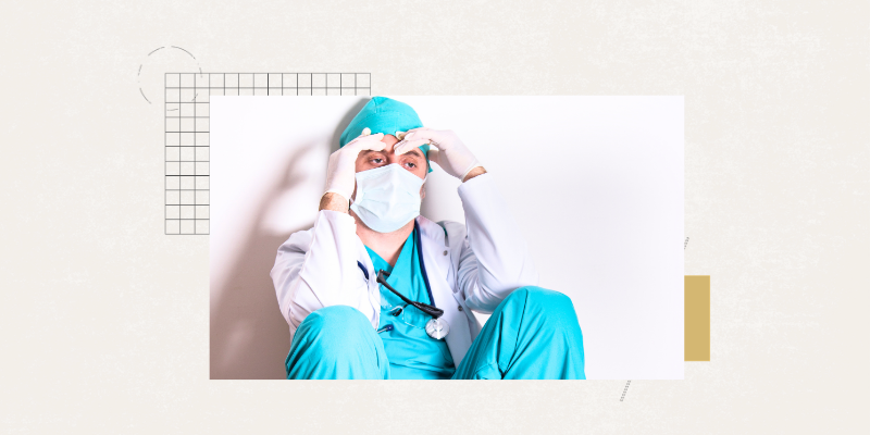 A tired-looking doctor wearing a mask and sitting against a wall